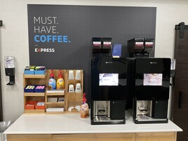 Una estación de café con dos máquinas de café, un dispensador de gel antibacterial y un cartel que dice 'NECESARIO. CAFÉ.'