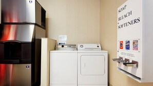 Un espace buanderie compact comprenant un réfrigérateur en acier inoxydable, un lave-linge et un sèche-linge. Un panneau affiché au mur indique les produits de lessive disponibles.