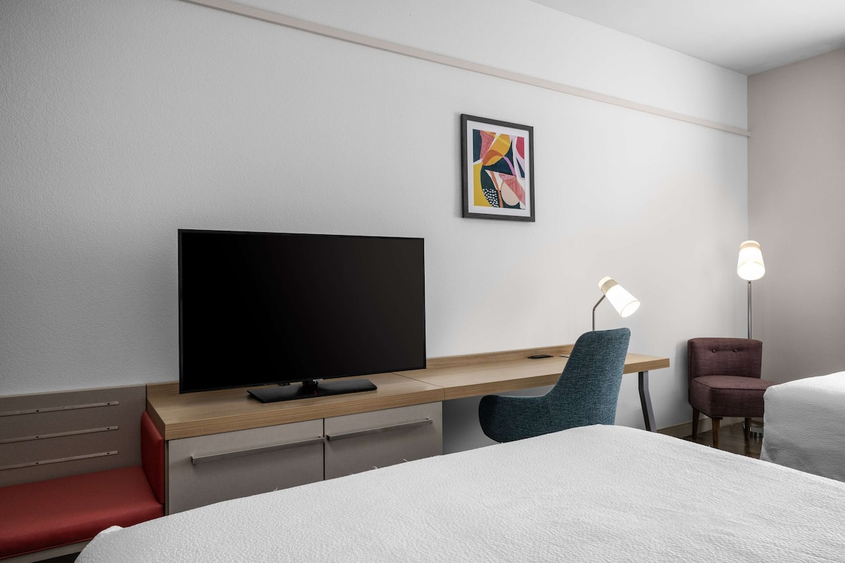 Ein modernes Hotelzimmer mit einem großen Flachbildfernseher, einem Schreibtisch mit Stuhl, einem Nachttisch und einer Lampe.