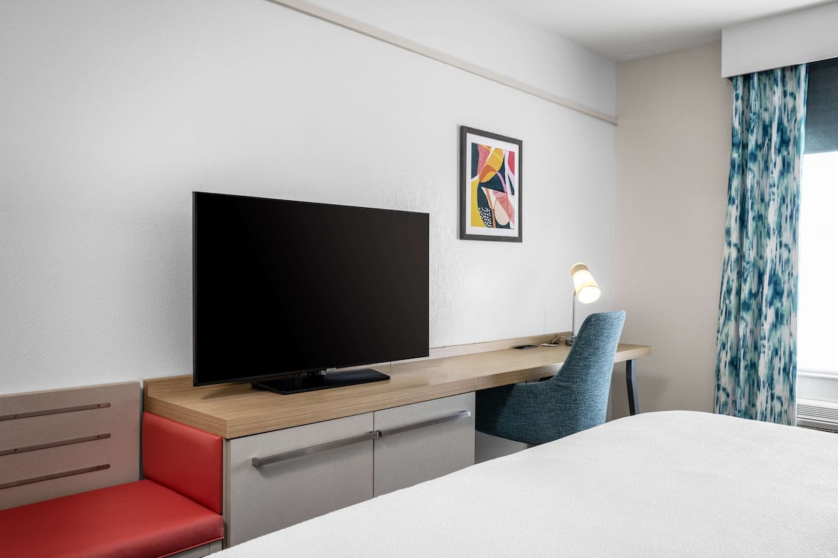 Ein Hotelzimmer mit einem Bett, einem Schreibtisch mit Fernseher, einem Sessel, einer Schreibtischlampe und einem Fenster mit Vorhängen.