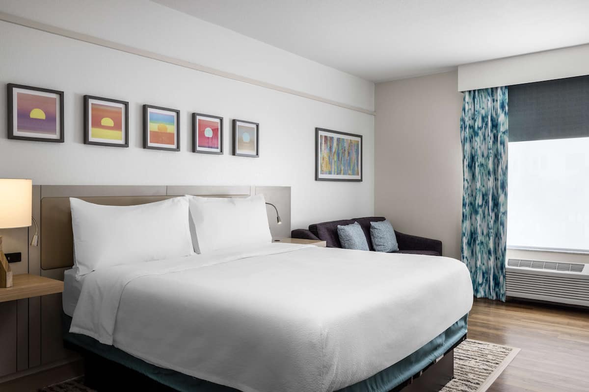 Ein Hotelzimmer mit einem großen Bett, einem Nachttisch, einem Schreibtisch, einem Sessel und einem Fenster mit Vorhängen.