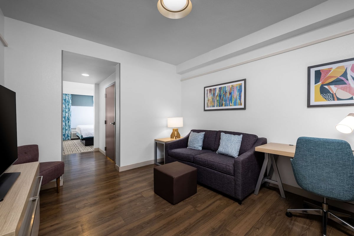 Ein modernes Wohnzimmer mit einem dunkelblauen Sofa, einem Holztisch, einem blauen Sessel und einem kleinen Schreibtisch mit Lampe. An der Wand hängt ein Gemälde und eine Tür führt in einen anderen Raum.