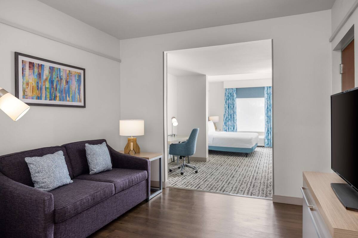 Ein modernes Hotelzimmer mit einem violetten Sofa, einem Schreibtisch mit Lampe, einem Fernseher und einem Bett mit einem blau-weißen Streifenmuster Vorhang.