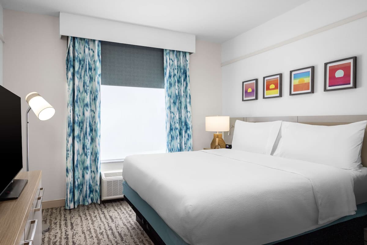 Ein Hotelzimmer mit einem Bett, einem Schreibtisch mit Lampe, einem Fernseher, einem Fenster mit Vorhängen und vier gerahmten Bildern an der Wand.
