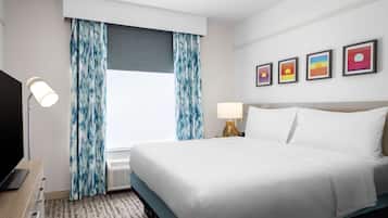 Une chambre d’hôtel avec un lit, un bureau avec une lampe, une télévision, une fenêtre avec des rideaux et quatre tableaux encadrés accrochés au mur.