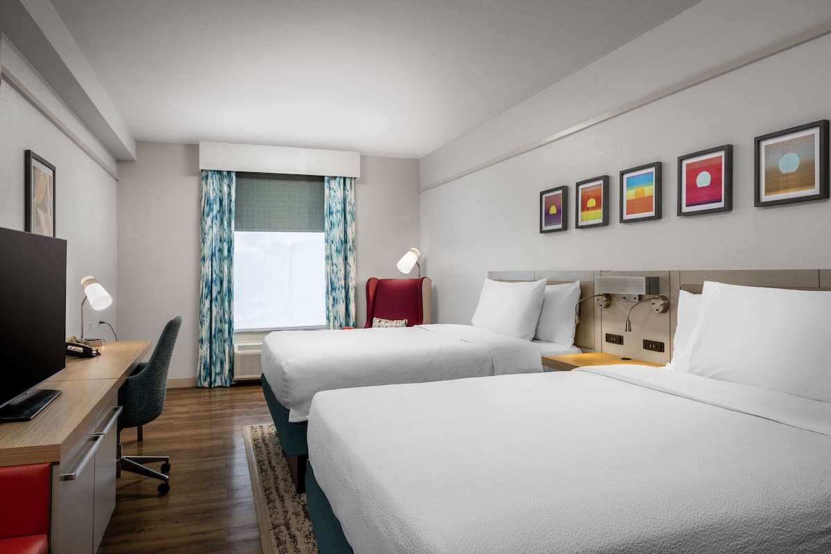 Ein Hotelzimmer mit zwei Betten, einem Schreibtisch mit Stuhl, einem Fernseher und einem Fenster mit Vorhängen.