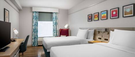 Ein Hotelzimmer mit zwei Betten, einem Schreibtisch mit Stuhl, einem Fernseher und einem Fenster mit Vorhängen.