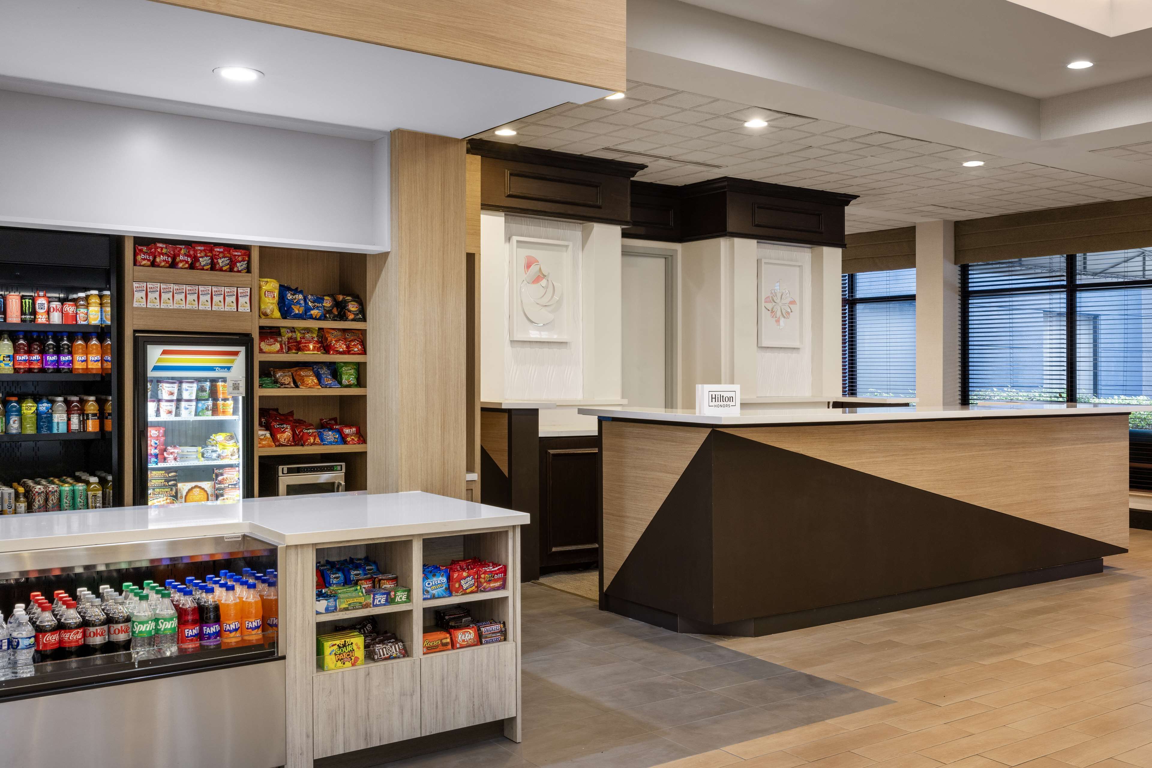 En hotellobby med en receptionsdisk, hyllor fyllda med snacks och drycker samt ett stort fönster som släpper in naturligt ljus.