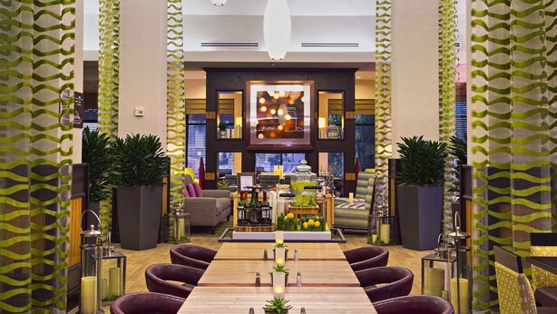 A modern hotel lobby with a long reception desk, comfortable seating, and a central bar area. - Hilton Garden Inn West Palm Beach Airport (West Palm Beach)