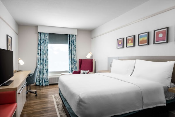 Una habitación de hotel con cama, escritorio con silla, televisión, sillón rojo y ventana con cortinas.
