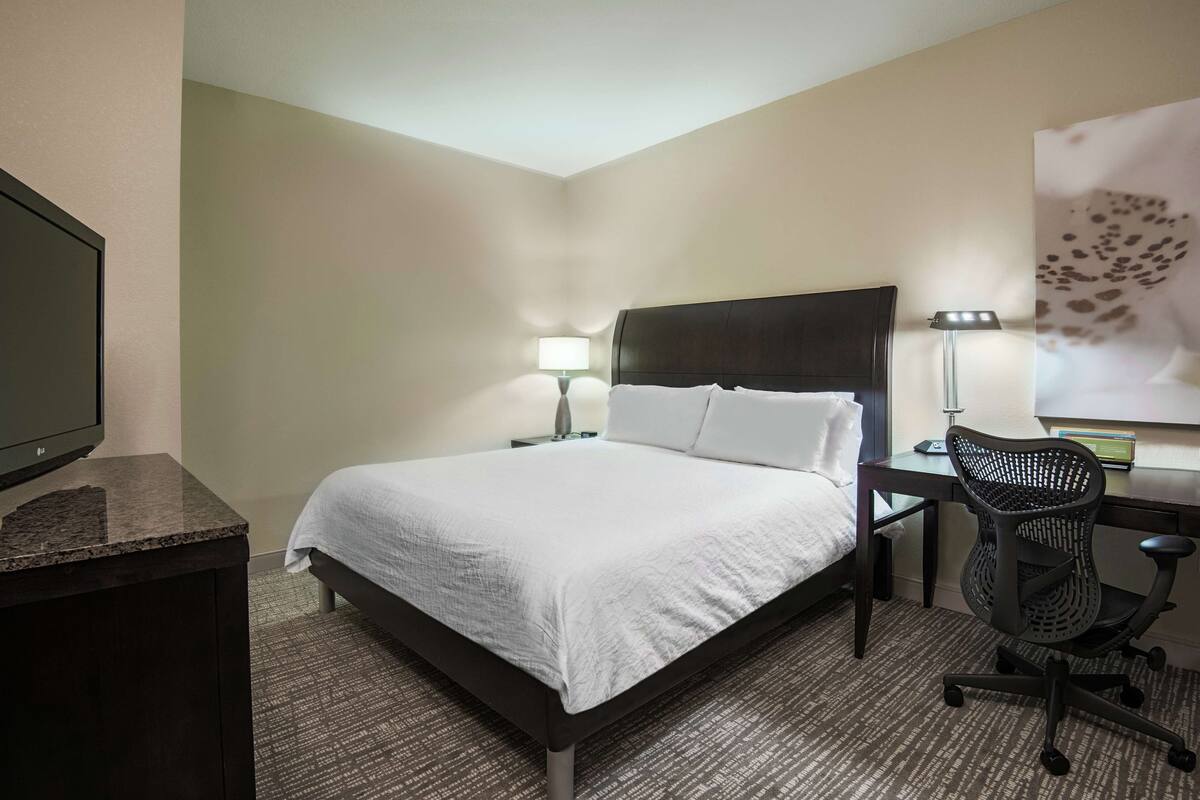 Ein Hotelzimmer mit Bett, Schreibtisch, Stuhl, Lampe und Fernseher.