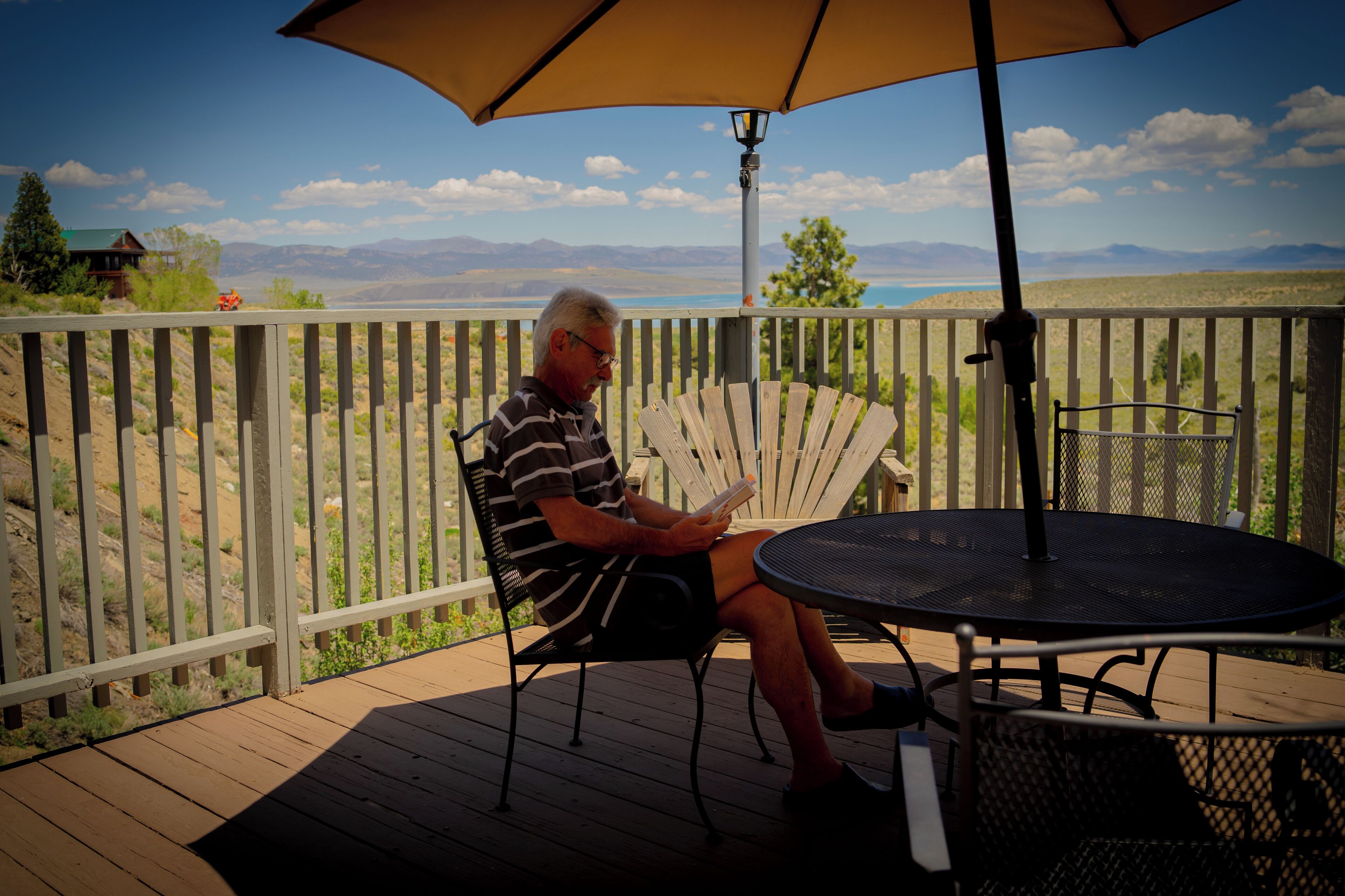 A man sitting on a deck chair reading a book, with a table and umbrella in the foreground and a scenic view in the background.