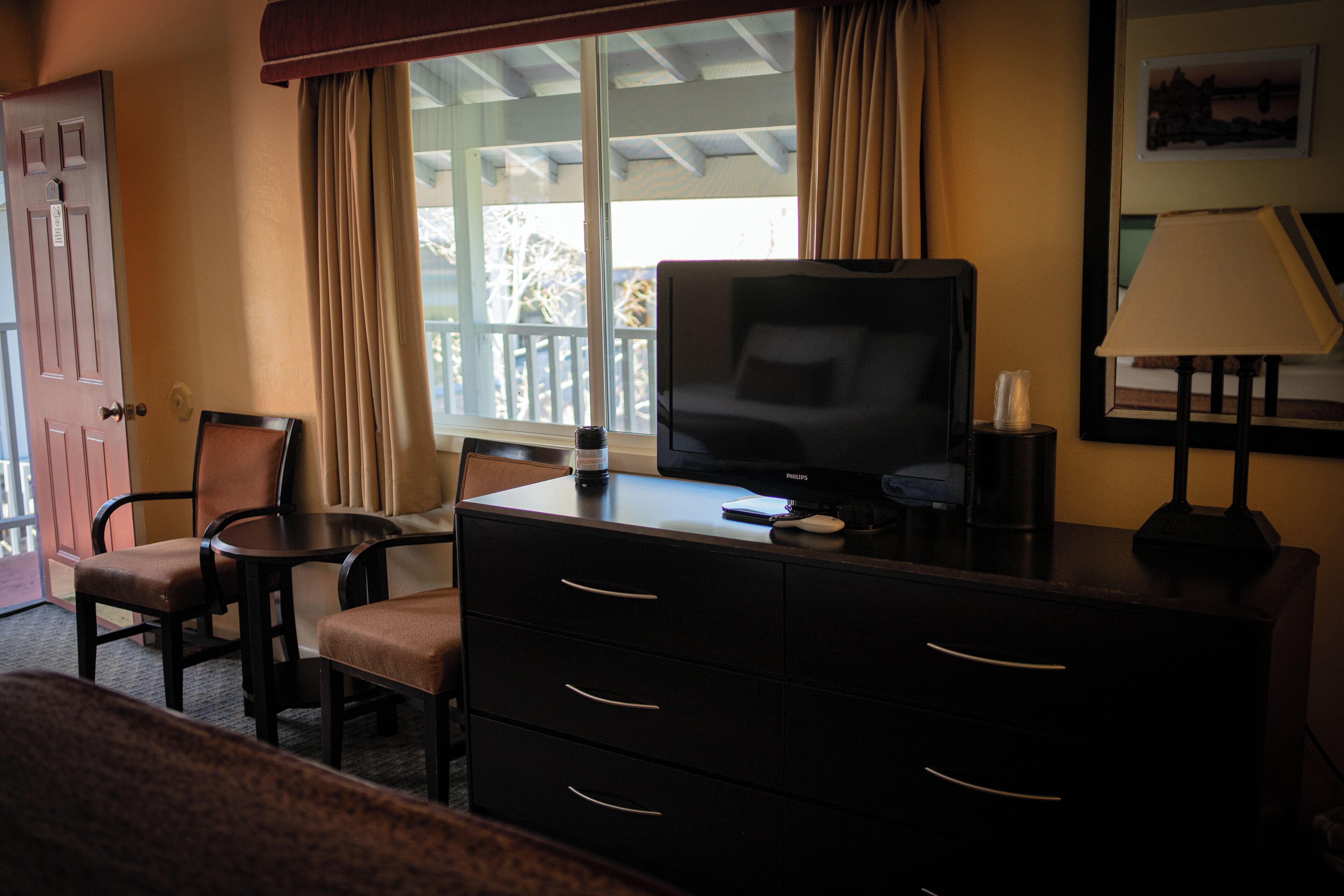 A hotel room with a television on a dresser, a chair, a desk, and a window with curtains.