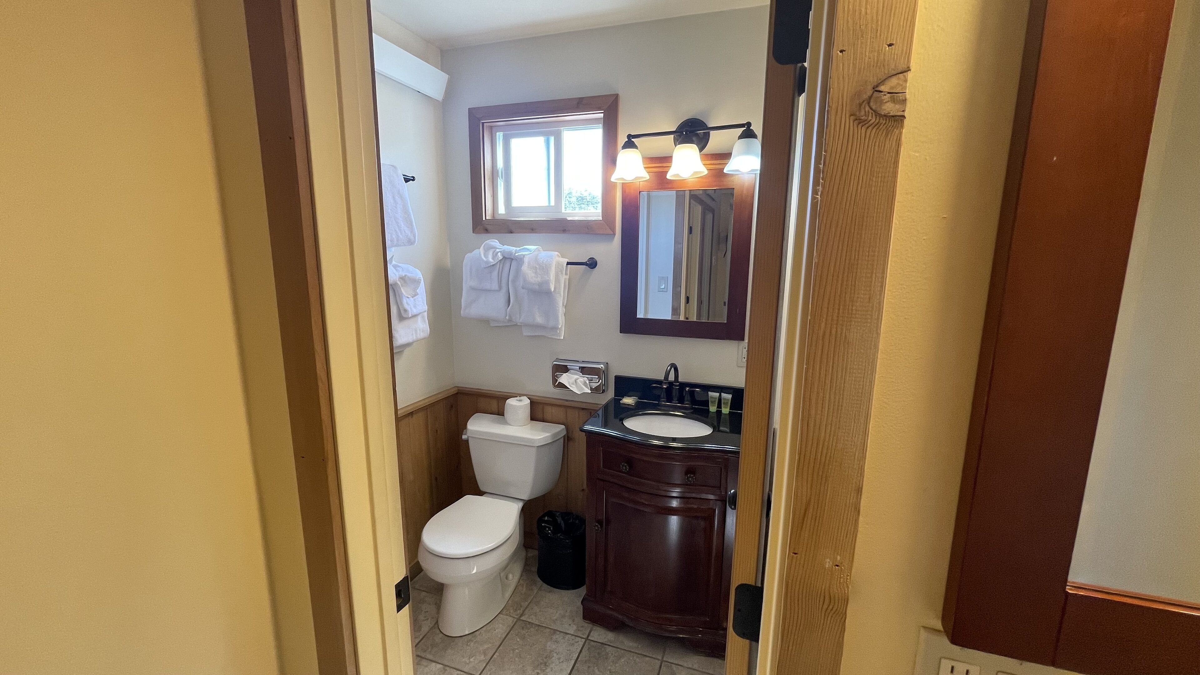 Ein Badezimmer mit Toilette, Waschbecken und Spiegel. Handtücher hängen an der Wand, und über der Toilette befindet sich ein Fenster.