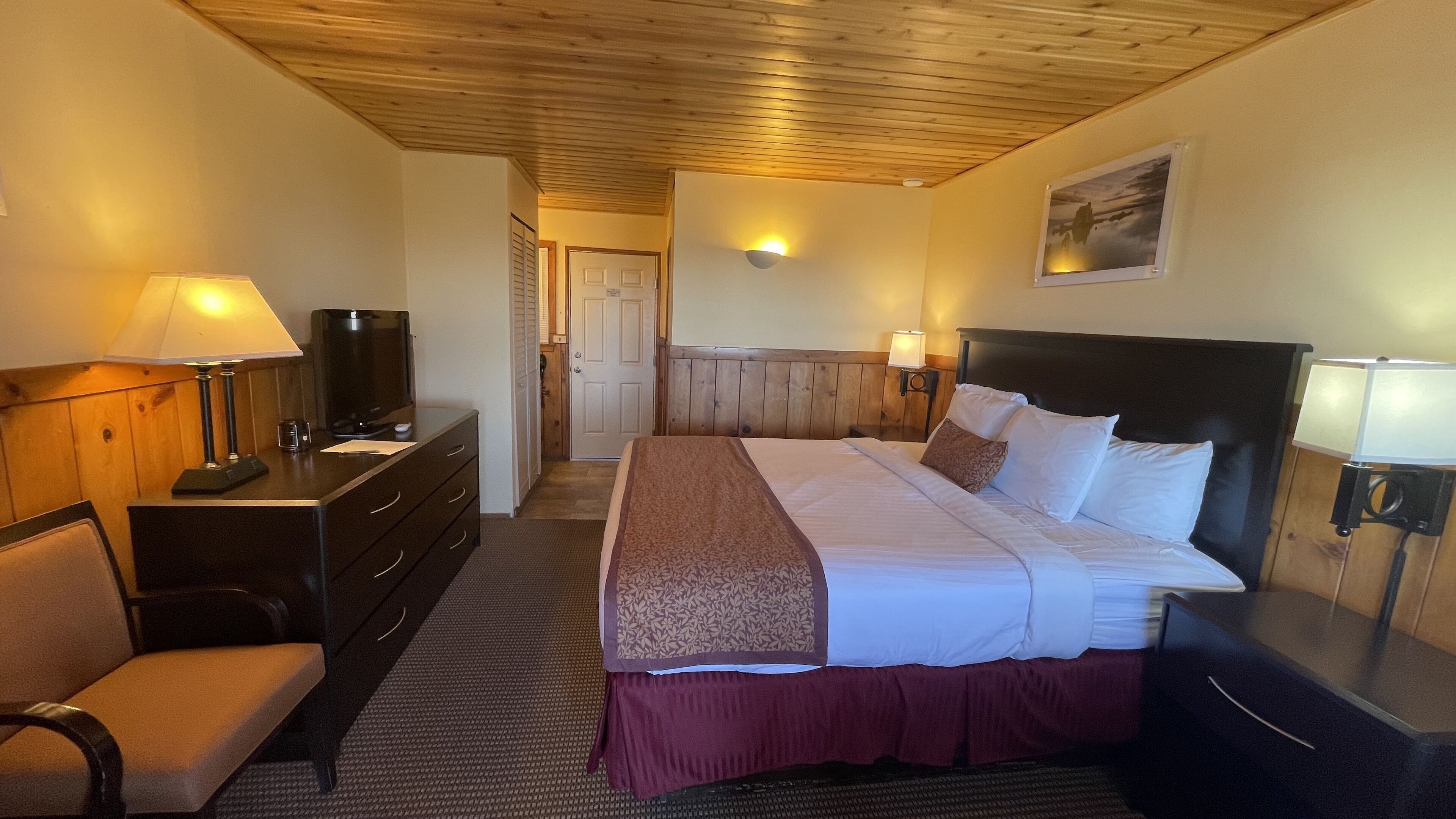 A hotel room with a wooden ceiling, a bed with white linens, a desk with a lamp, a TV, and a chair.