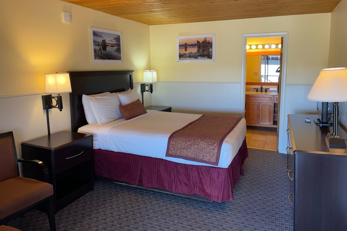 Habitación de hotel con cama, dos mesitas de noche con lámparas, una silla, un escritorio y un espejo. En la pared, se observan dos cuadros enmarcados.
