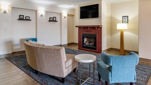 Un salon avec un canapé beige, un fauteuil bleu, une cheminée, un téléviseur fixé au mur et une lampe.