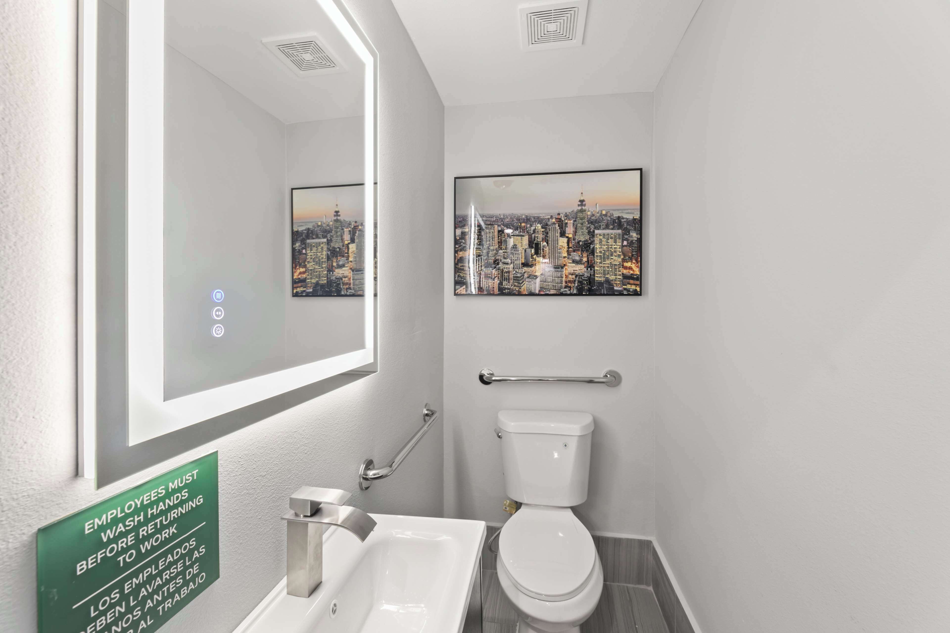 Un baño moderno con una pintura de paisaje urbano, dispensador de gel antibacterial y un letrero que indica a los empleados que se laven las manos antes de volver al trabajo.