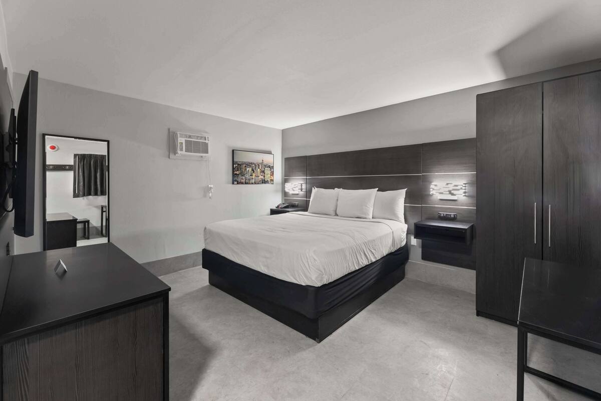 A modern bedroom with a large bed, a dark wooden wardrobe, a bedside table, and a wall-mounted air conditioning unit.