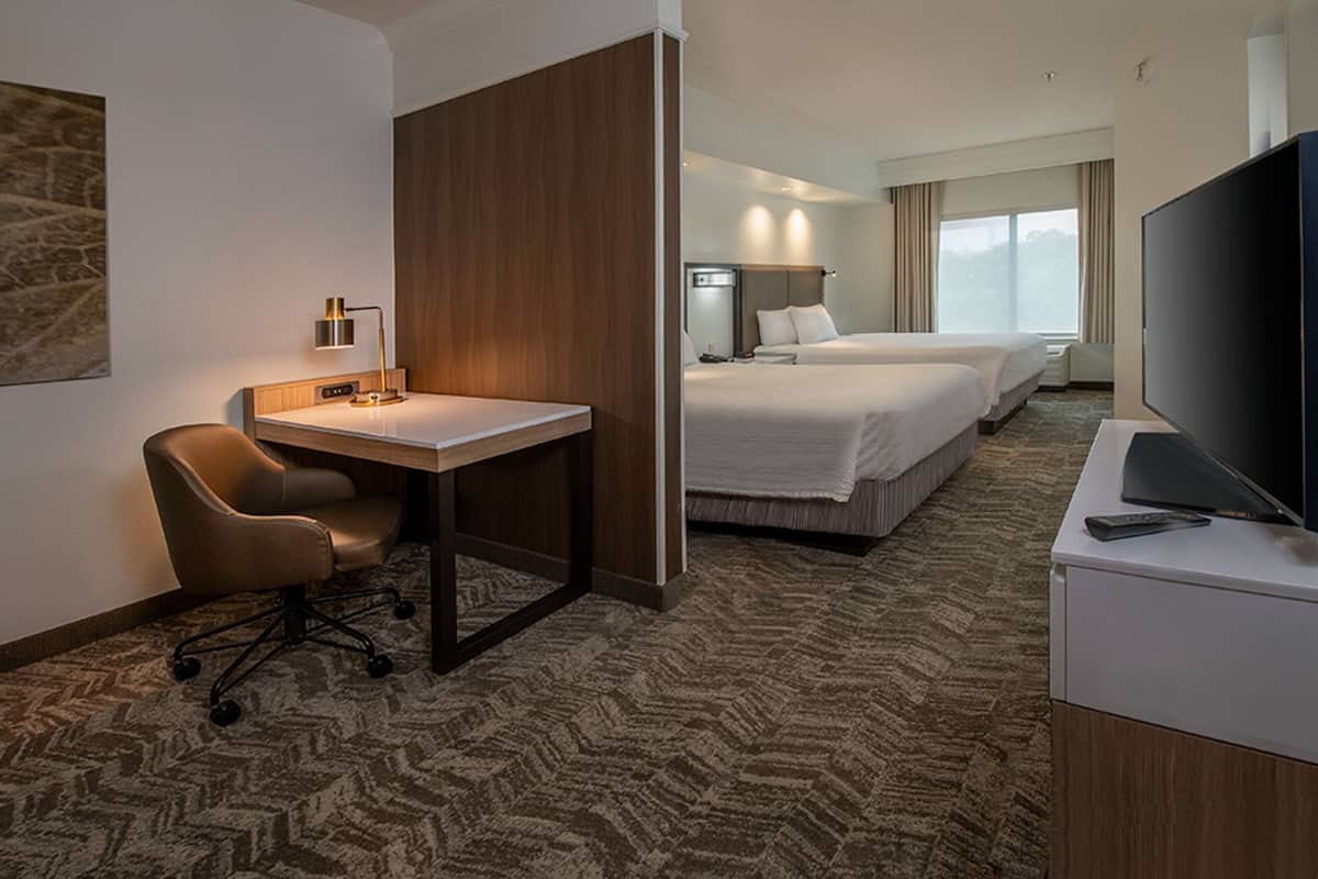 Habitación de hotel con escritorio, silla, dos camas, televisión y un ventanal grande.