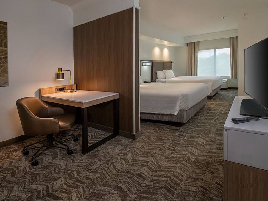 Een hotelkamer met een bureau, stoel, twee bedden, een televisie en een groot raam.