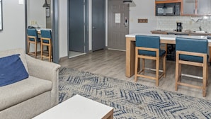 Ein modernes Wohnzimmer mit einer Couch, einem Couchtisch und einem gemusterten Teppich. Es gibt einen Essbereich mit Tisch und Stühlen sowie eine Küche mit Mikrowelle und Schränken.