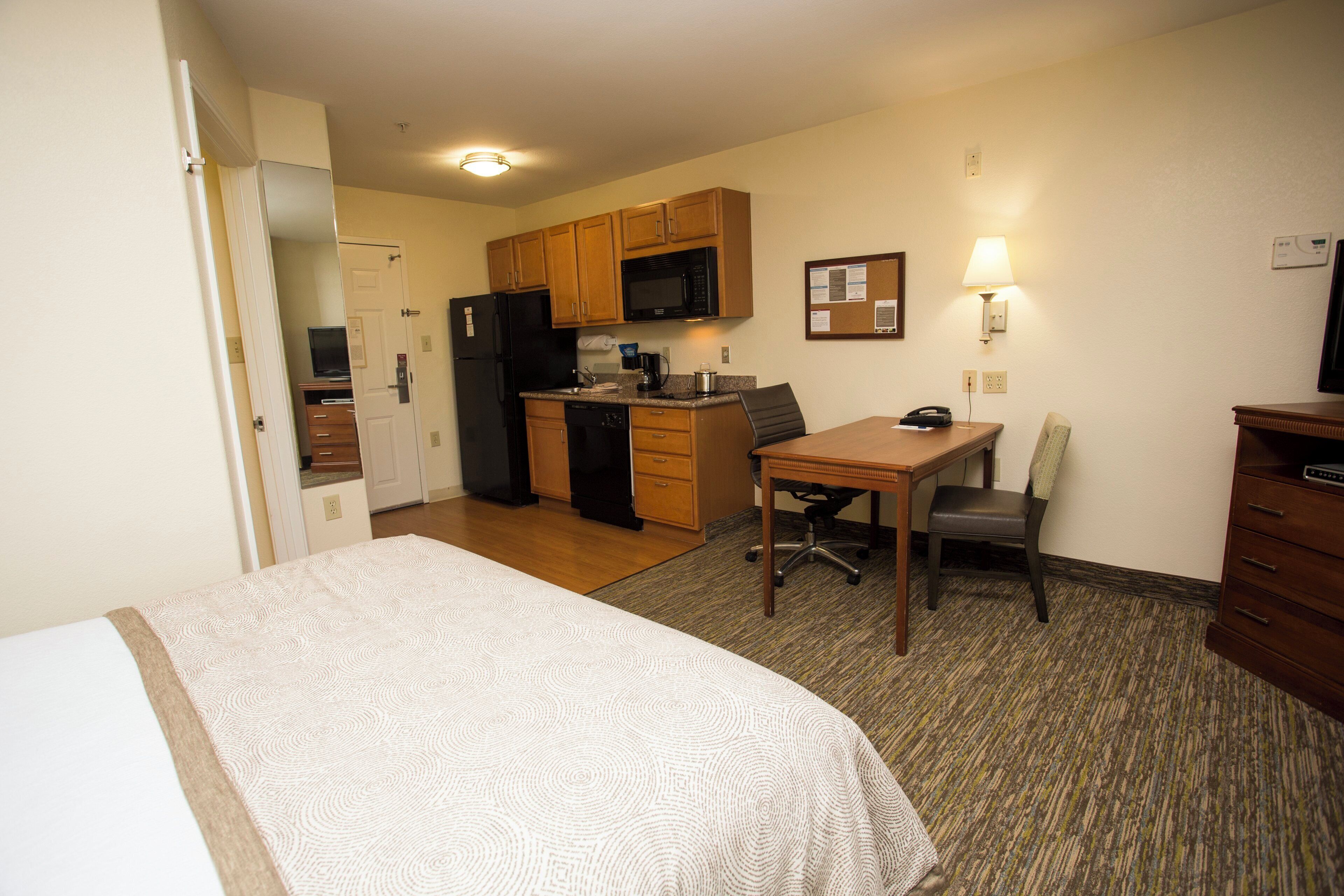 A compact hotel room with a bed, a desk, a chair, a kitchenette, and a small dining area.