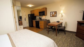 A compact hotel room with a bed, a desk, a chair, a kitchenette, and a small dining area.