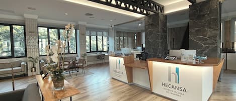 A modern hotel reception area with a wooden reception desk, a seating area with a sofa and chairs, and a large window offering a view of the outdoors.