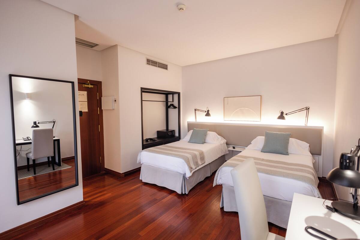 A hotel room with two beds, a desk, a chair, a mirror, and a bedside lamp.
