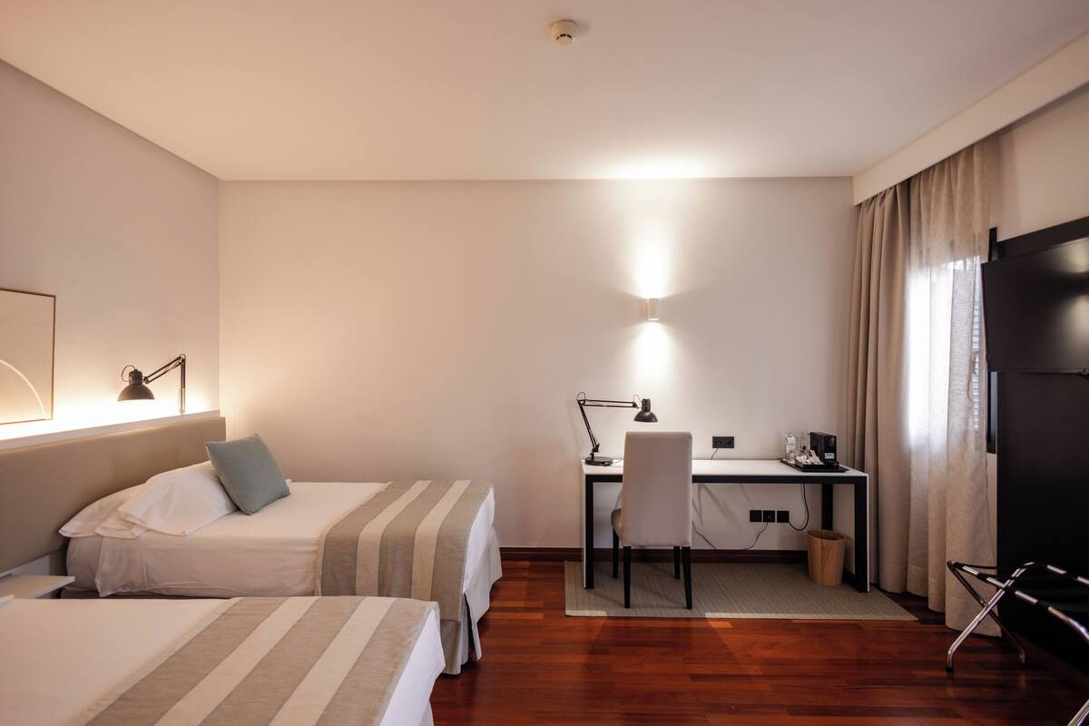 A hotel room with two beds, a desk, a chair, a TV, and a lamp.