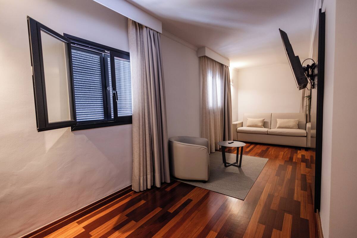 A modern living room with a sofa, a small table, a chair, a window with blinds, and a television mounted on the wall.