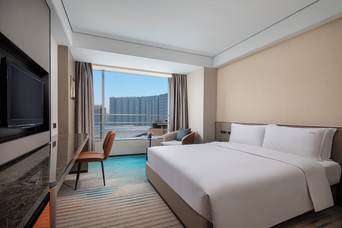 A hotel room with a large bed, a TV, a desk with a chair, and a view of a cityscape.