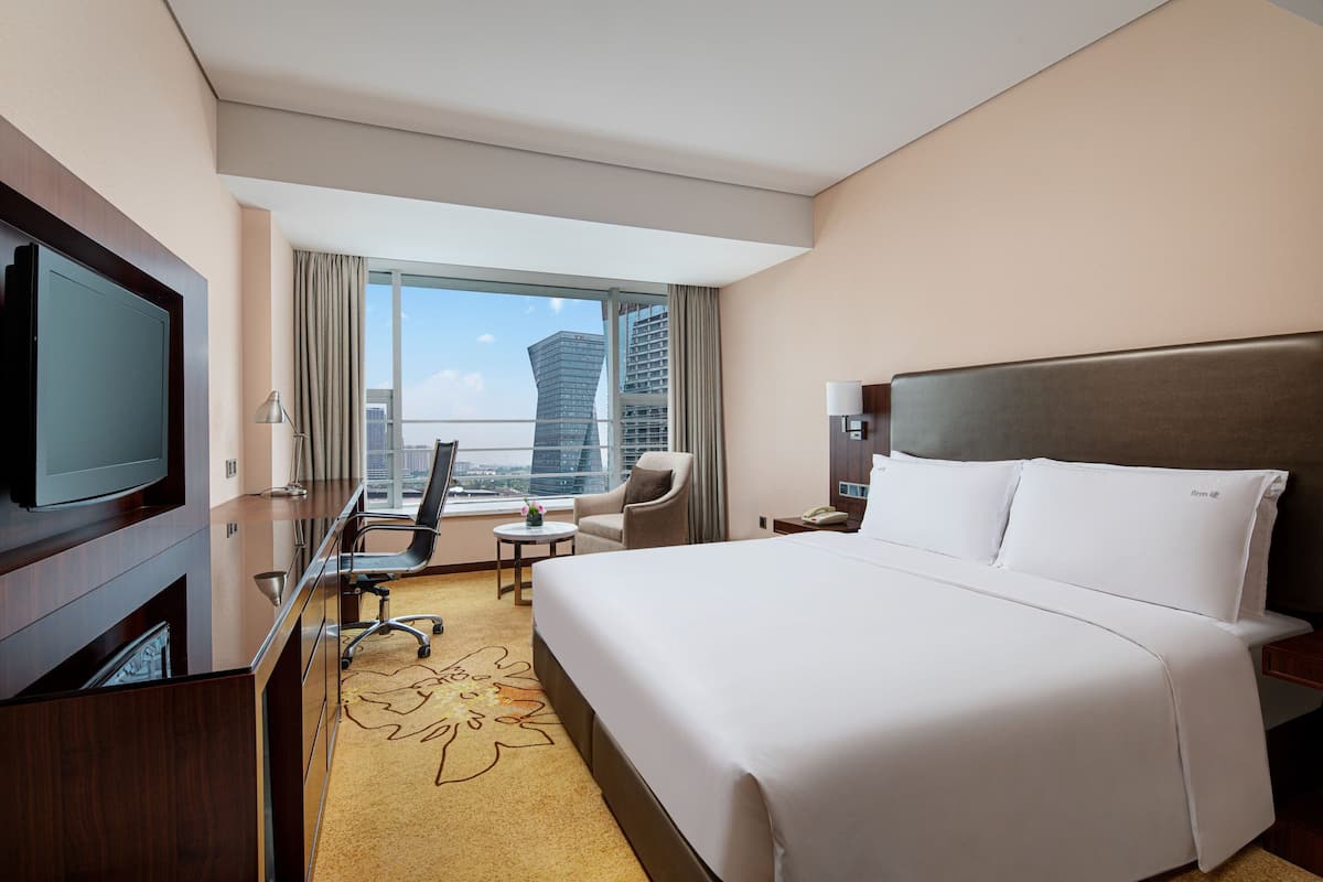 A hotel room with a large bed, a desk with a flat-screen TV, a chair, a small table, and a view of the cityscape through large windows.