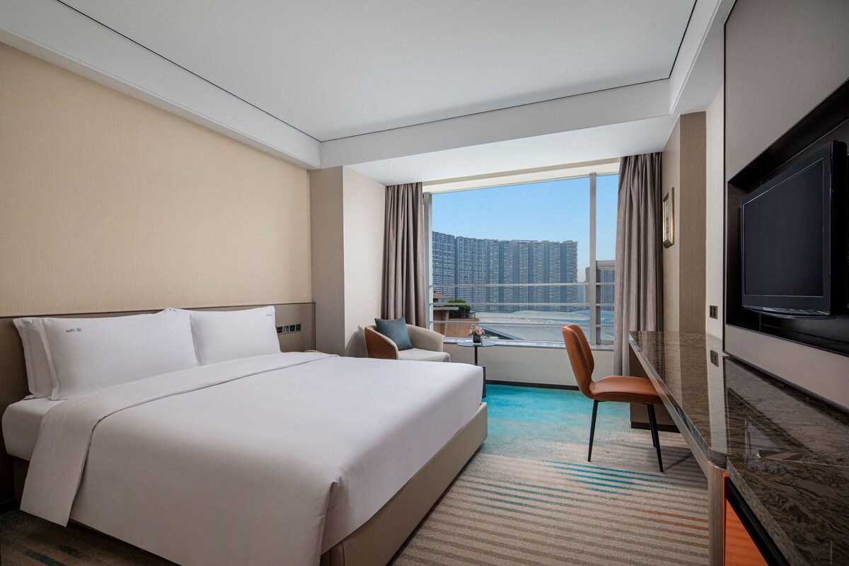 A hotel room with a large bed, a TV, a desk, and a view of high-rise buildings.