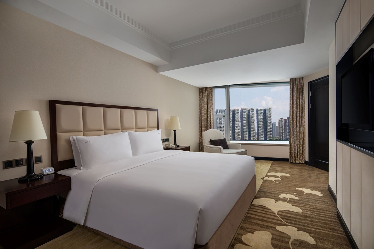 A hotel room with a large bed, two bedside tables with lamps, a TV, and a view of the city skyline.
