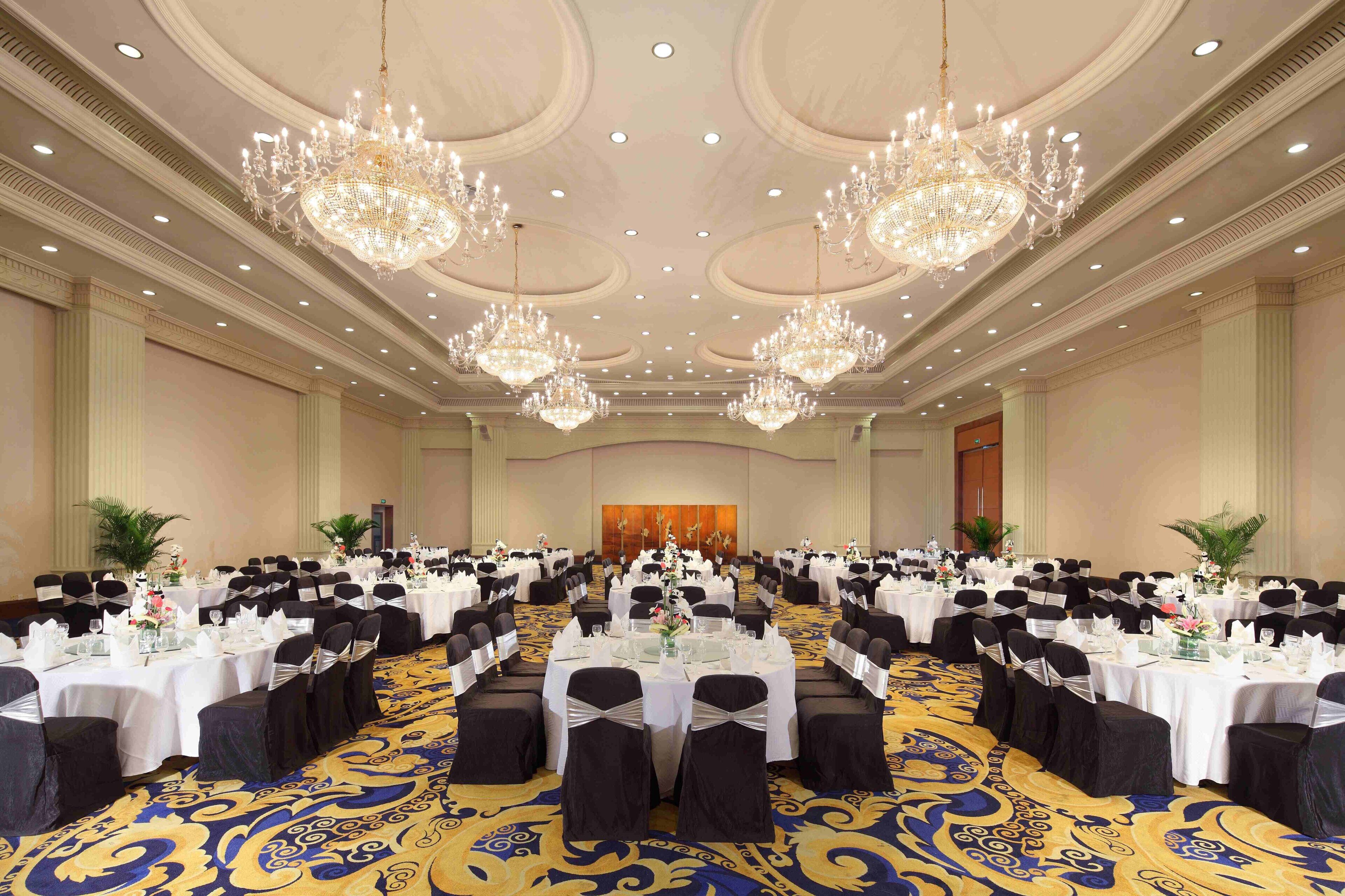 A large banquet hall with multiple round tables, black chairs, and chandeliers.