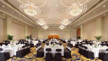 A large banquet hall with multiple round tables, black chairs, and chandeliers.
