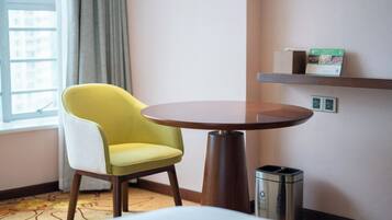Habitación de hotel con una silla amarilla, una mesa redonda, un cubo de basura, una estantería con una caja y una ventana con cortinas.