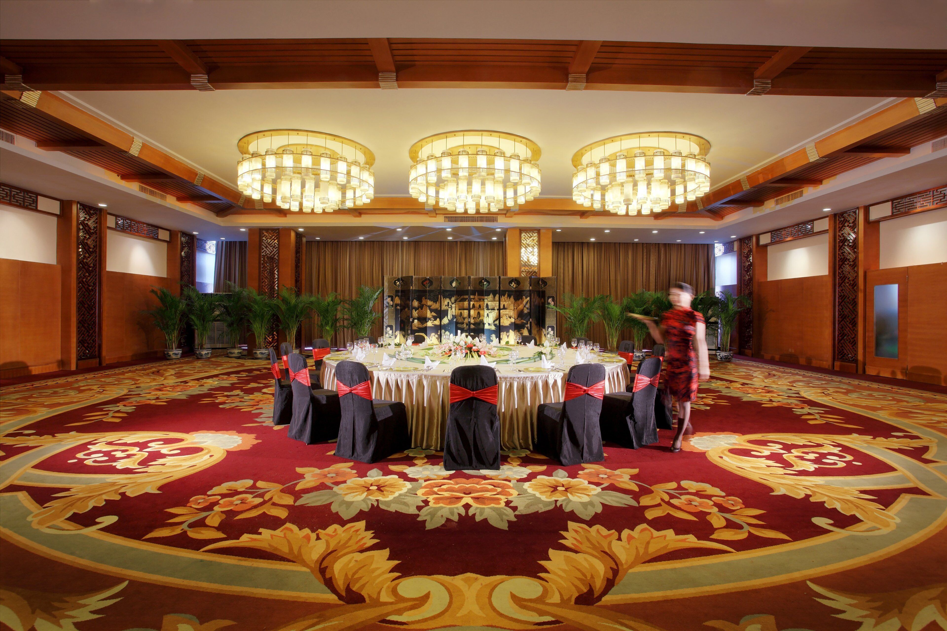 A banquet hall with a round table set for a formal event, surrounded by chairs with red sashes, and a large floral carpet.