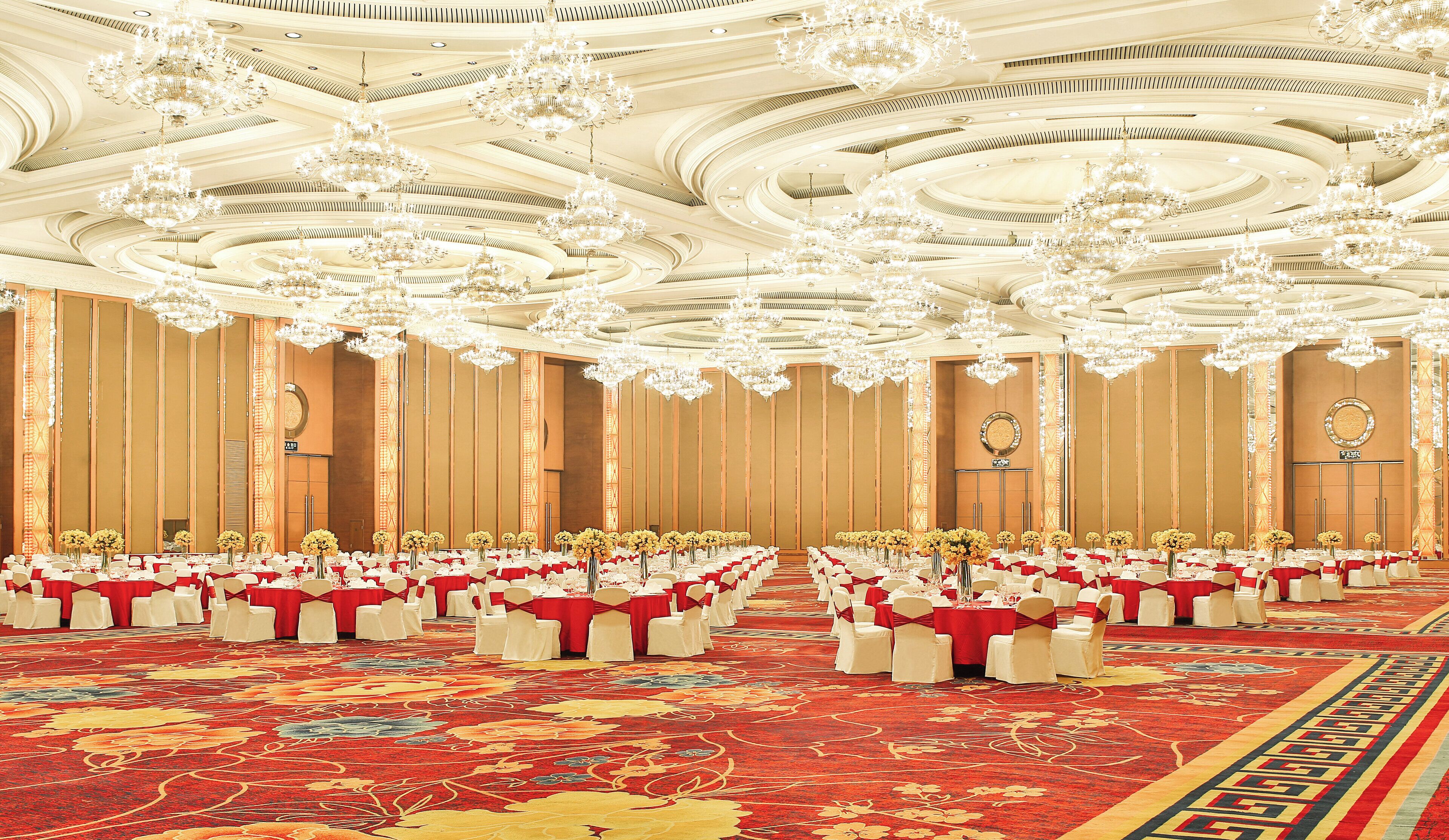 Un amplio salón de banquetes con filas de mesas, sillas blancas y manteles rojos.