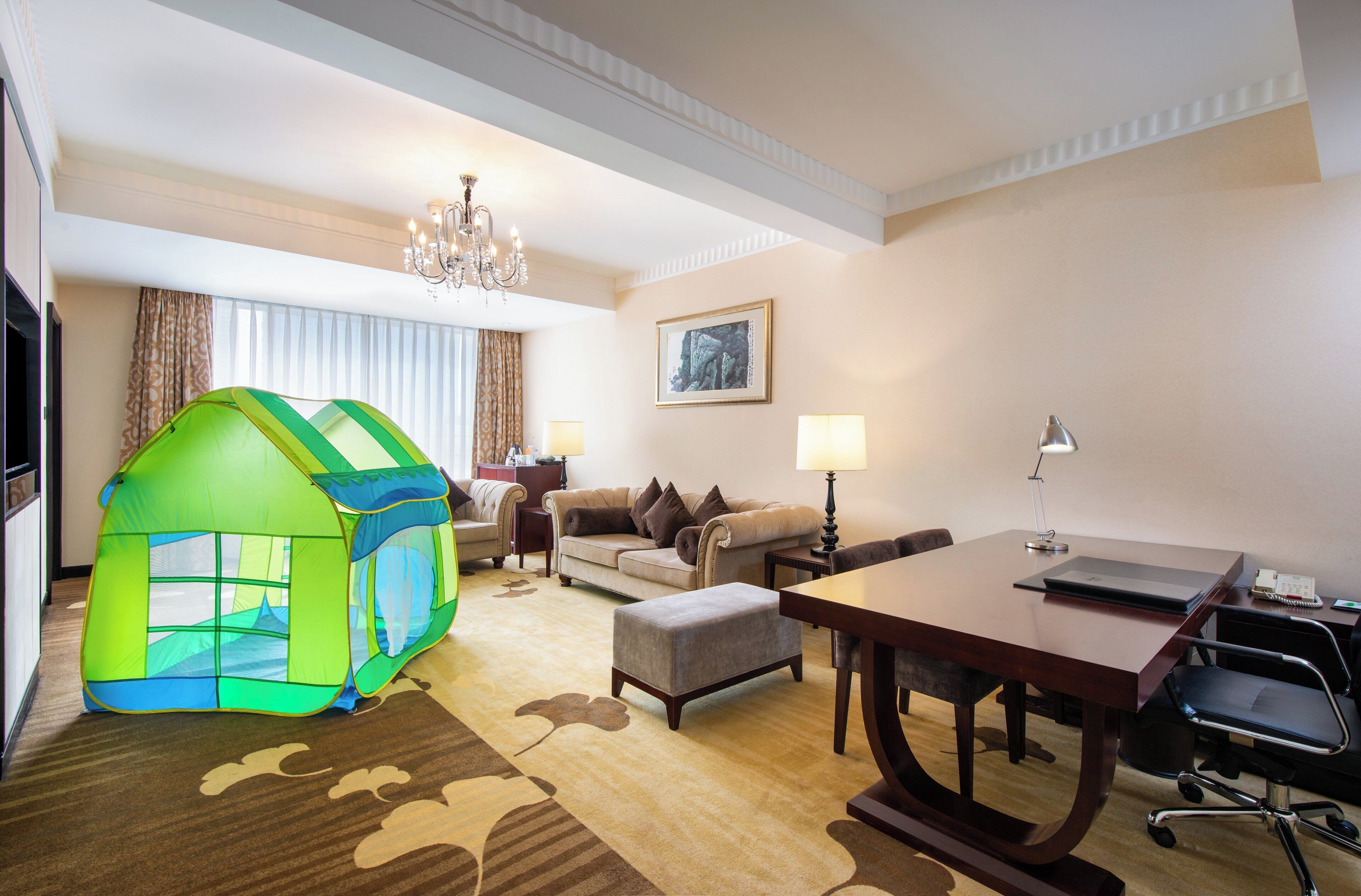 A hotel room with a large bed, a desk, a chair, a lamp, a chandelier, and a green play tent.