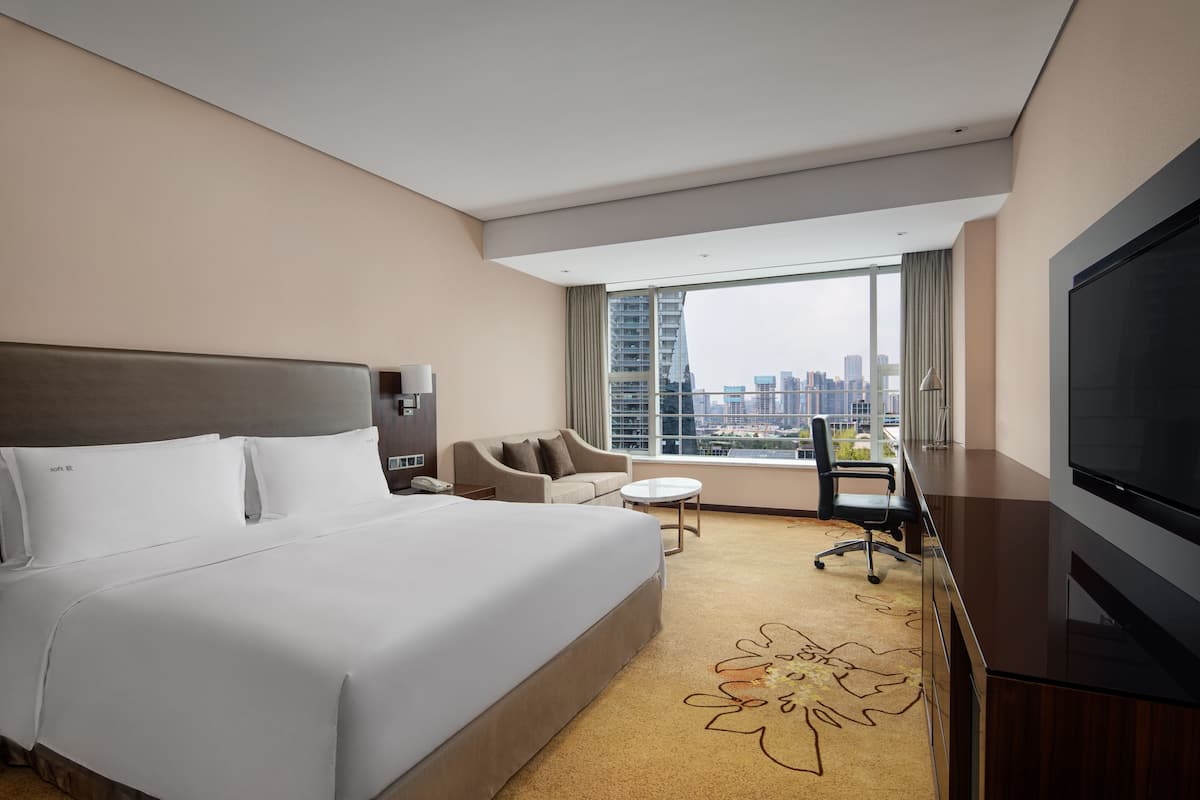 A hotel room with a large bed, a desk with a TV, a sofa, a small table, and a view of the city.