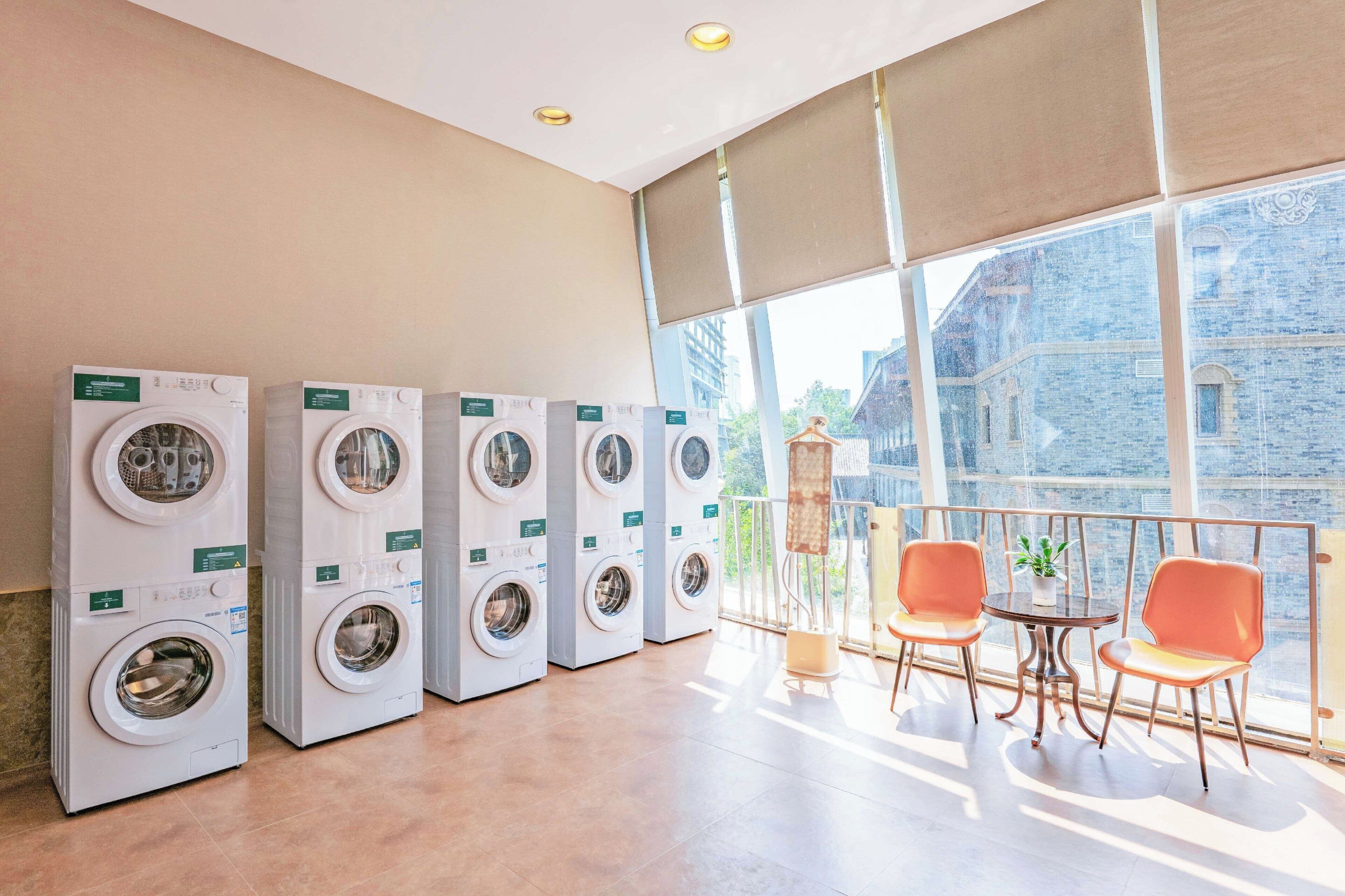 A laundry room with multiple washing machines, a small table with chairs, and a large window with a view of buildings.