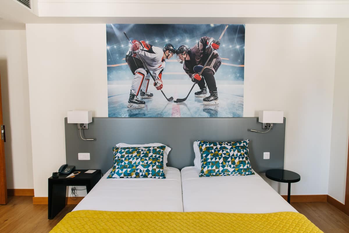 A hotel room with a bed, two patterned pillows, a bedside table with a phone, a small table with a lamp, and a large sports-themed wall mural.
