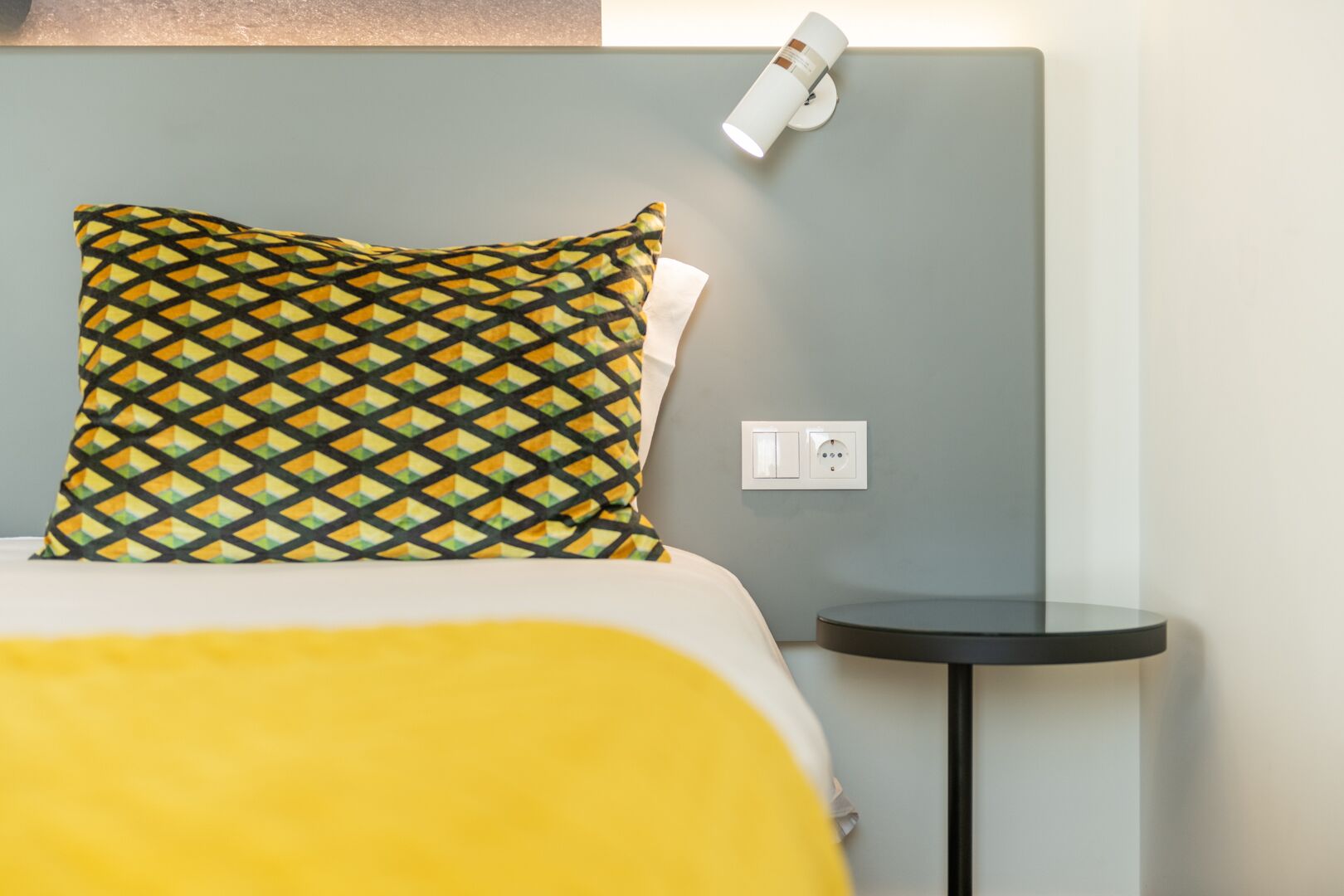 A bed with a patterned pillow, a bedside table, and a wall-mounted light fixture.