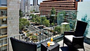 Un balcón con mobiliario de mimbre, barandilla de vidrio y vista a una calle de la ciudad con edificios y coches estacionados.