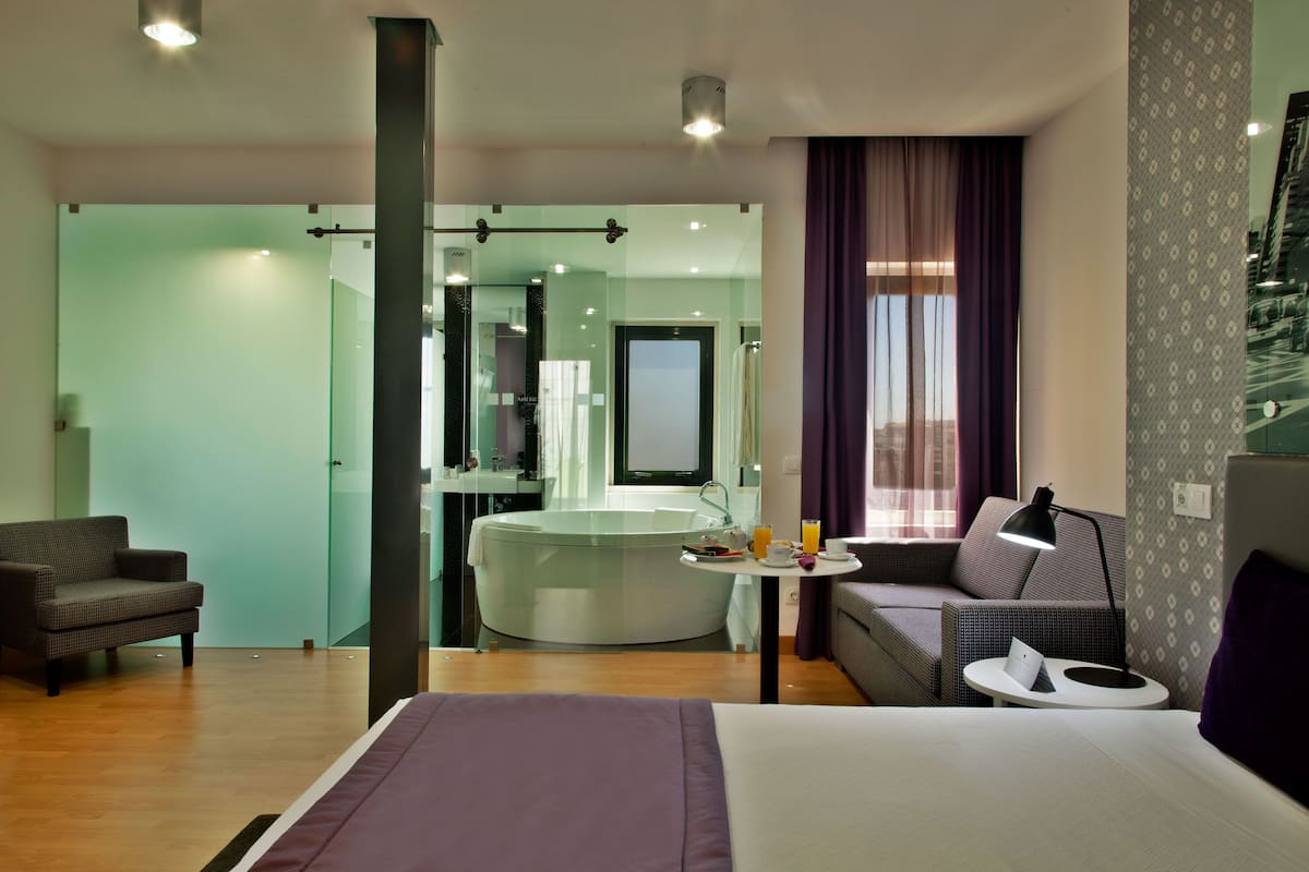 A modern hotel room with a large bed, a sitting area with a sofa and armchair, a small table with a lamp, and a glass-enclosed bathroom with a bathtub.