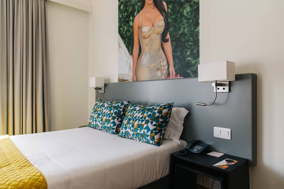 A hotel room with a bed, patterned pillows, a nightstand with a phone, and a painting on the wall.