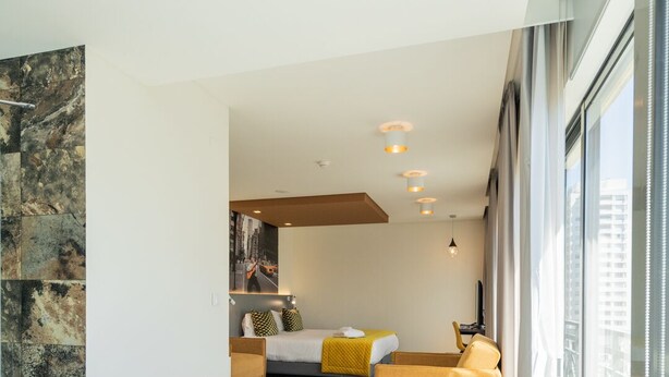 Habitación de hotel moderna con una bañera grande, una cama con ropa de cama amarilla y vistas a la ciudad a través de amplios ventanales.